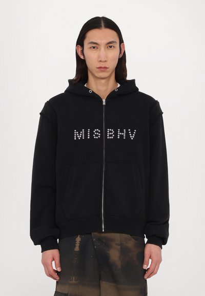 Black zip-up hoodie with a hood, featuring "MISBHV" in small silver studs across the chest and front kangaroo pockets. Textured fleece material.