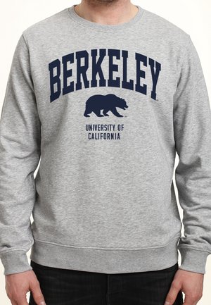 Mann trägt ein hellgraues Sweatshirt mit der Aufschrift "BERKELEY" und einem Bärenlogo über dem Text "University of California" in dunkelblauer Schrift.