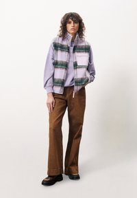 Weste mit gestreiftem Muster in pastelllila und grün, kombiniert mit einem helllila Sweatshirt und braunen, weit geschnittenen Cordhosen. Schwarze Schuhe.