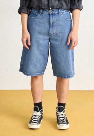 Personne portant un short en jean bleu jusqu'aux genoux, des baskets montantes noires avec des lacets blancs, des chaussettes noires et une chemise à carreaux foncée avec les manches retroussées.