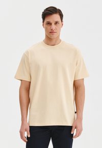 Jasno beżowa bawełniana koszulka z okrągłym dekoltem i krótkimi rękawami. Posiada luźny krój i minimalistyczny design bez widocznych logotypów czy wzorów.