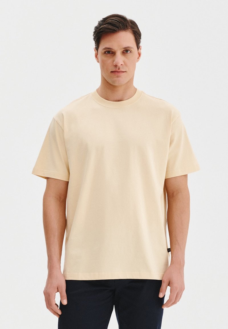 Jasno beżowa bawełniana koszulka z okrągłym dekoltem i krótkimi rękawami. Posiada luźny krój i minimalistyczny design bez widocznych logotypów czy wzorów.