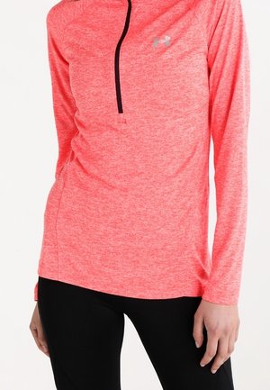 Femme portant un pull sportif corail rose à manches longues avec une fermeture éclair noire et un pantalon noir, debout avec une main détendue sur le côté.
