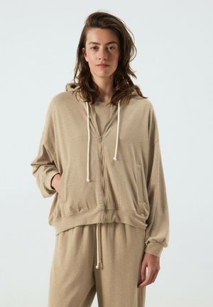 Donna con capelli castani di media lunghezza che indossa una felpa beige con zip e pantaloni beige abbinati con coulisse, in piedi con una mano in tasca.