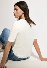T-shirt blanche à manches courtes avec un col rond, fabriquée en tissu léger, associée à un jean en denim bleu, en pose assise contre un fond clair.