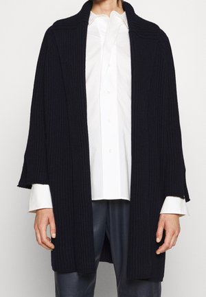 Person trägt eine schwarze, offen vorne getragene, gerippte Strickjacke über einem weißen Hemd mit Knöpfen und schwarzen Hosen vor einem einfarbigen weißen Hintergrund.