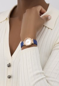 Roosa kuldne käekell ümmarguse näo, valge numbrialuse, sinise silikoonriba ja teravate aktsentidega. Kätel, mis on valge soonikutega sviidri vastu.