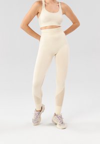 Un reggiseno sportivo beige chiaro e leggings a vita alta con pannelli in rete testurizzati. Il tessuto sembra elastico, con polsini aderenti alla caviglia e scarpe da ginnastica.