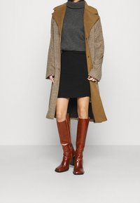 Kollad brun kappa med en stor krage över en grå polotröja, kombinerad med en svart minikjol och knähöga blanka bruna stövlar.