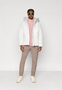 Frau, die eine weiße Daunenjacke, ein rosa Zip-up-Oberteil, beige Jogginghosen, weiße Sneaker und Sonnenbrille trägt, vor einem schlichten weißen Hintergrund steht.