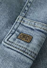 Šviesiai mėlynas džinsinis audinys su matomomis audimo linijomis, su siuvinėtų lopu su tekstu ir kontrastingu auksinės spalvos siūlu išilgai siūlių.