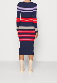 Abito midi a coste con strisce orizzontali nei colori blu navy, rosso, grigio chiaro e viola. Maniche lunghe e silhouette aderente. Indossato con stivali alla caviglia color crema.