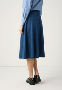 Frau in einem blauen Midi-Denimrock, gestreiftem blauen Hemd und schwarzen klobigen Loafers, die vor einem einfarbigen Hintergrund steht.