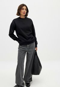 Sudadera negra con un cuello alto, combinada con jeans de pierna ancha grises. El modelo lleva un gran bolso tote negro y usa zapatos negros.