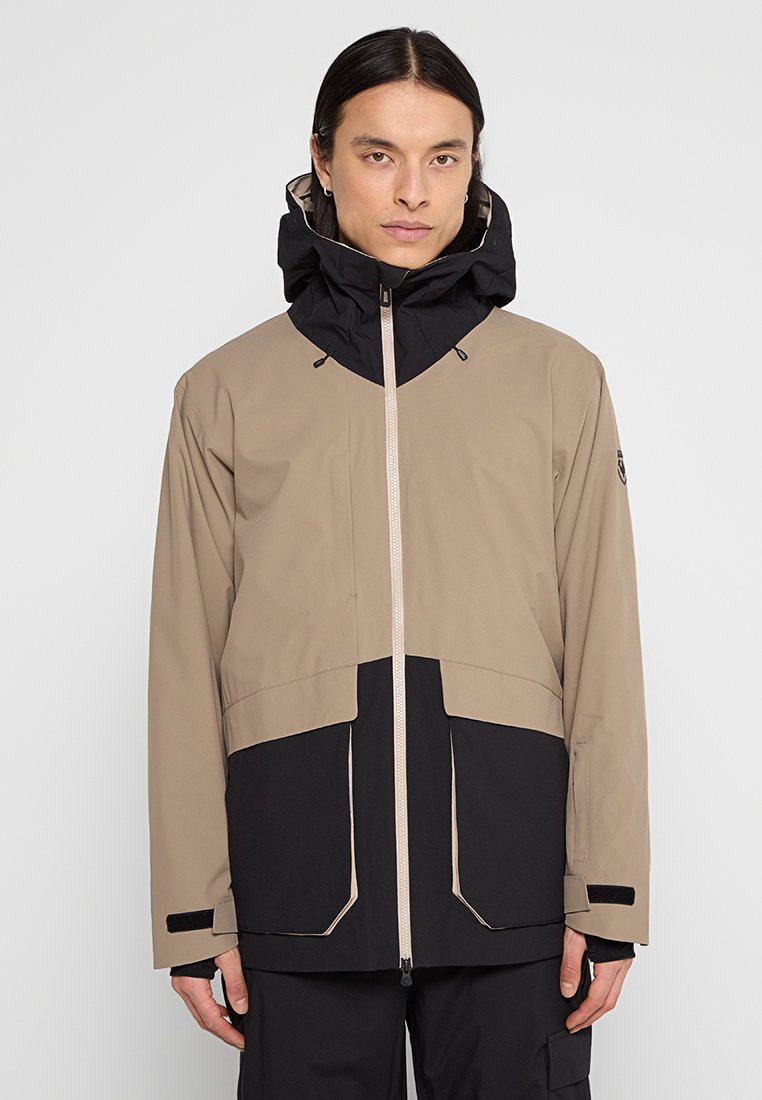 Beige und schwarze wasserdichte Jacke mit hohem Kragen, verstellbarem Kapuze und zwei vorderen Taschen. Verfügt über einen durchgehenden Reißverschluss und Klettverschluss-Bündchen.