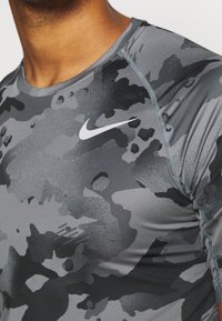 Grå camouflageträningströja i stretchmaterial, med rund hals, långa ärmar och en vit Nike-logotyp på bröstet.