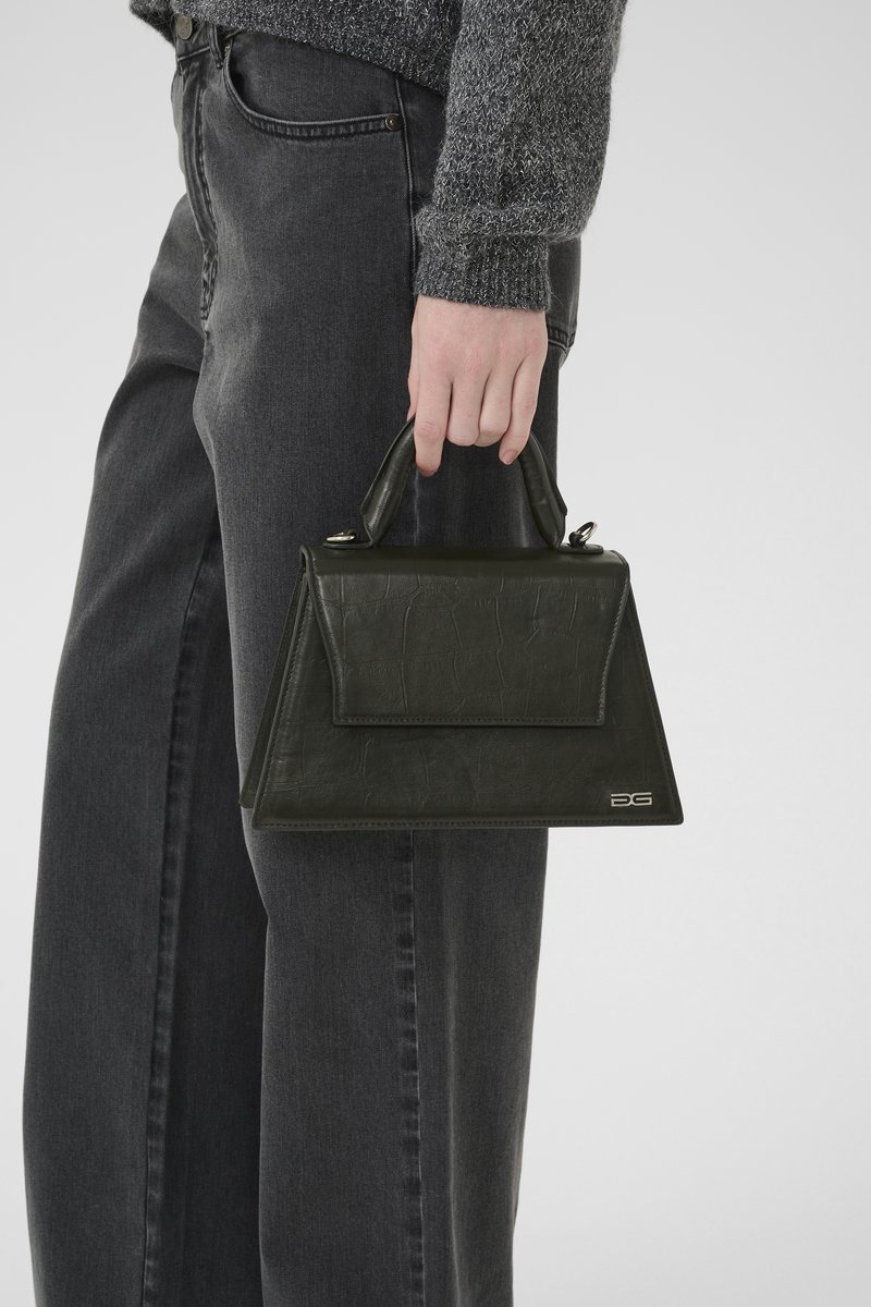 Person som bär en mörkgrå tröja och jeans, håller en liten mörkgrön texturerad handväska med topphandtag och silverlogotyp.