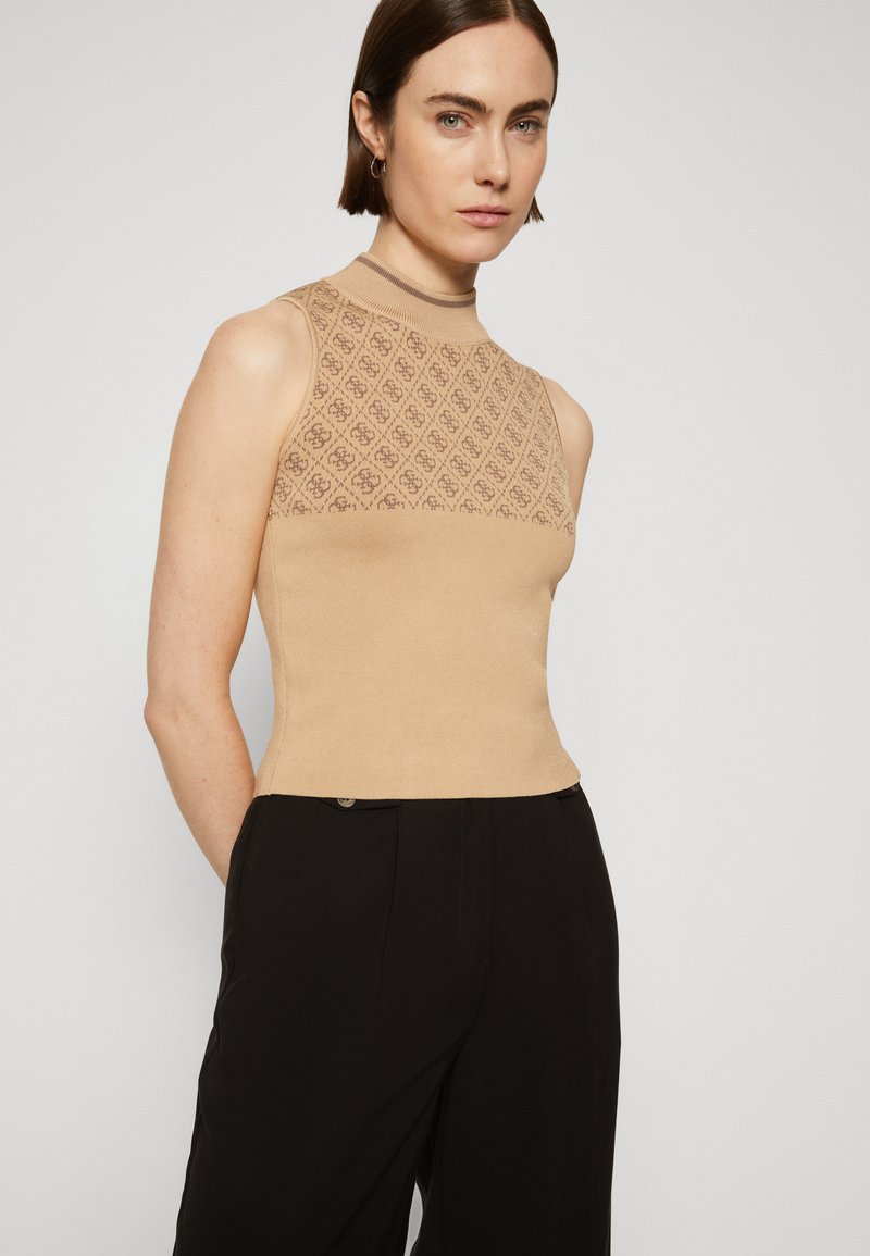 Maglione senza maniche beige con una parte superiore decorata e un colletto a costine, abbinato a pantaloni neri, su uno sfondo neutro.
