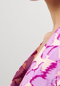 Livlig lilla og pink mønstret stof, der viser et flydende design med en V-hals og buet skulderlinje, som fremhæver en let tekstur.