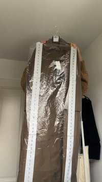 Borsa per abiti marrone che contiene un lungo cappotto marrone. Il cappotto è racchiuso in plastica trasparente con etichette bianche contrassegnate con "S" lungo i lati.