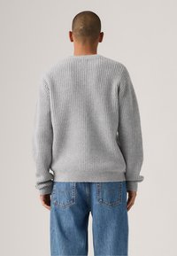 Grå strikkegenser med et teksturert mønster, rund hals og ribbestrikkede mansjetter. Brukt med lys blå denimjeans. Utsikt fra baksiden.