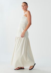 Jasno-beżowa sukienka maxi z prążkowanym, bezramiennym topem, zaznaczoną talią i rozkloszowanym dołem. Noszona z czarnymi płaskimi sandałami, o gładkiej teksturze.