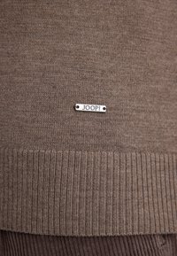 Brązowy sweter robiony na drutach z prążkowanym dołem i srebrną metką z logo "JOOP!" w dolnym lewym rogu. Miękka tekstura, casualowy styl.