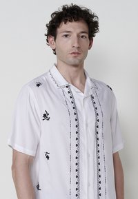 Camisa blanca de manga corta con bordado floral negro, detalles verticales y cierre de botones en la parte delantera. Tejido suave y ligero.