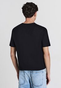 Uomo con capelli ricci e scuri, che indossa una maglietta nera a maniche corte senza fronzoli e jeans in denim blu chiaro, girato di spalle contro uno sfondo semplice.