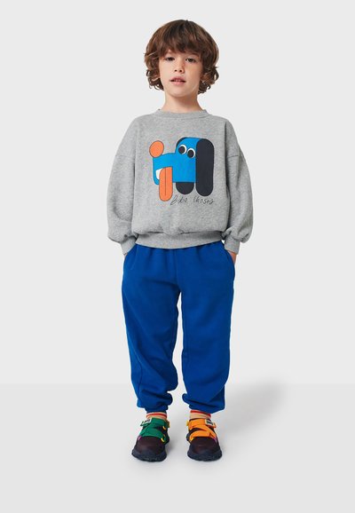 Felpa grigia con un grafico astratto colorato, abbinata a pantaloni blu. Le scarpe hanno fasce multicolori e una suola scura.