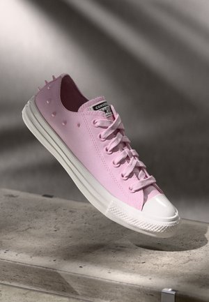Rosa canvas-sneakers med vit gummikappa på tårna, snörning och små upphöjda piggar längs sidorna; platt vit sula. Har en varumärkesetikett på tungan.