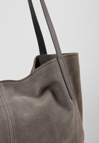 Sac fourre-tout en daim gris avec des anses en cuir lisse et des coutures visibles sur un fond clair uni.