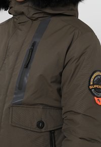 Olivengrøn dunjakke med hætte, med en sort lodret stribe, lynlåslomme og en orange logo-patch på ærmet.