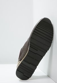 Sola di una scarpa Michael Kors marrone inclinata contro un muro bianco, che mostra il battistrada nero testurizzato e la incisione del nome del marchio.