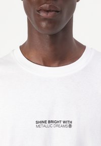 T-shirt bianco in cotone con scollo rotondo, decorato con testo stampato in nero che recita "BRILLA FORTE CON METALLIC DREAMS®" sulla parte anteriore.