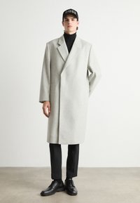 Abrigo de lana gris claro con cuello muesca, diseño de doble botonadura, combinado con un jersey de cuello alto negro, pantalones y zapatos negros, complementado con una gorra.