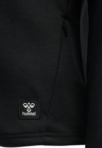 Pullover nero con tasca laterale con zip, tessuto morbido e etichetta logo bianca cucita con un design geometrico.
