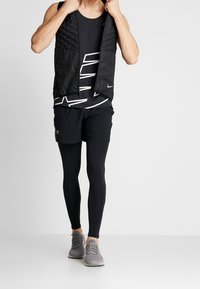 Czarna ocieplana kamizelka na zamek, założona na czarny top na ramiączkach z białym wzorem graficznym, w połączeniu z czarnymi szortami i legginsami.