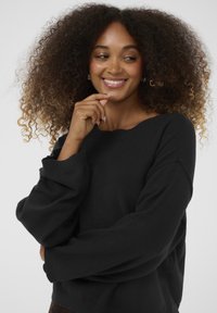 Crni oversized pleteni pulover s širokim izrezom i dugim, labavim rukavima. Glatka tekstura i opušteni kroj, u kombinaciji s neutralnom pozadinom.