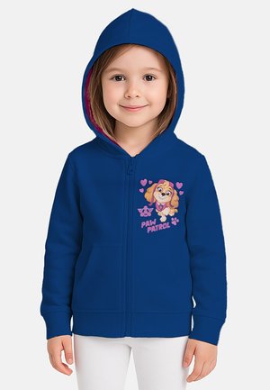 Mädchen in blauer Kapuzenjacke mit Paw Patrol Charakter und pinken Herzen auf der Brust, kombiniert mit weißen Hosen, lächelt nach vorne.