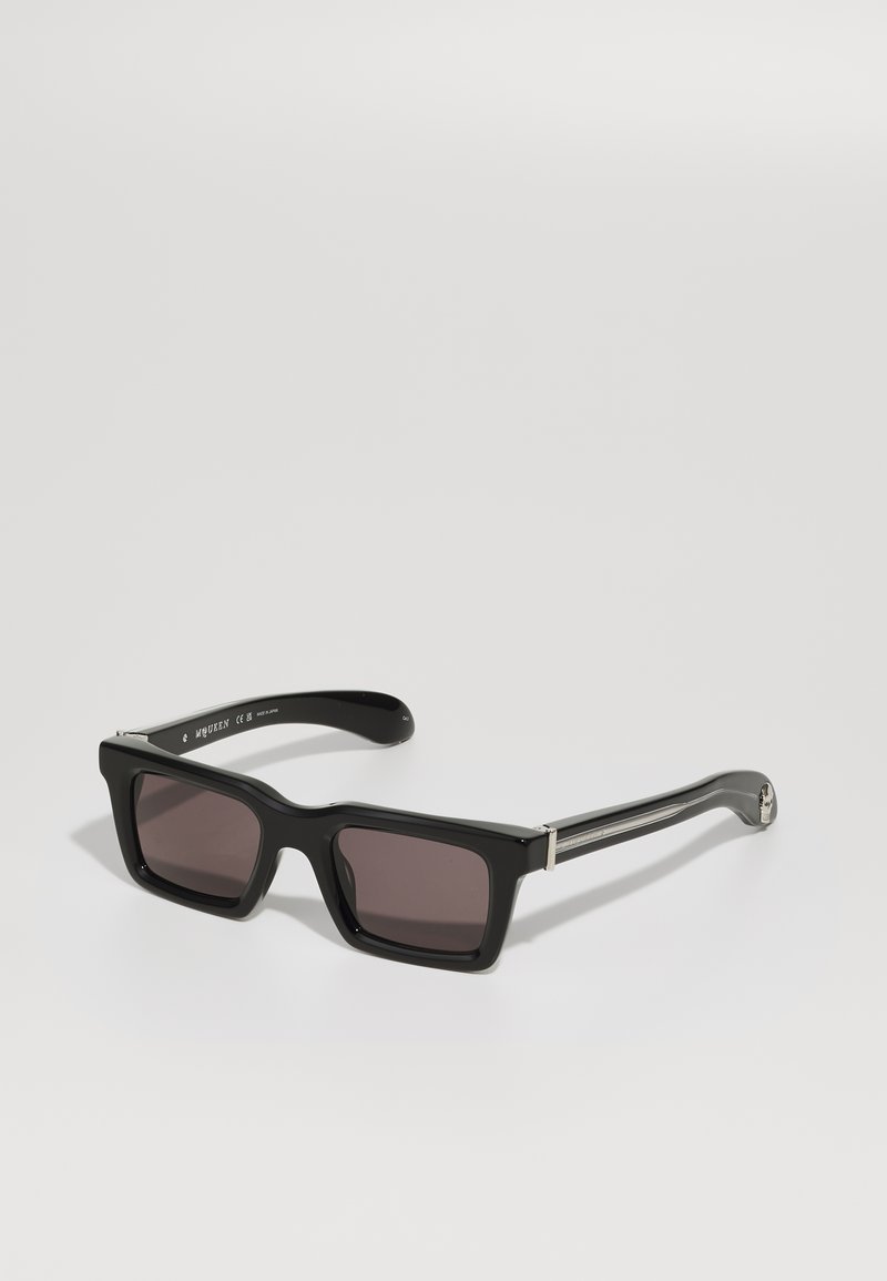 Gafas de sol rectangulares negras con lentes oscuros, que presentan montura de plástico brillante y detalles en tono plateado en las patillas.