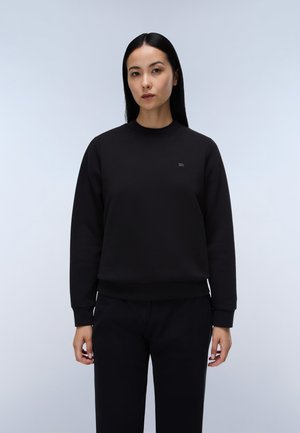 Kvinde med lige sort hår iført en ensfarvet sort sweatshirt og sorte bukser, står foran en lysegrå baggrund.