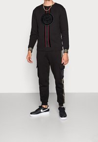 Felpa nera con logo centrale, strisce verticali rosse e nere. Abbinata a pantaloni joggers neri con tasche laterali e dettagli dorati. Sneakers nere.