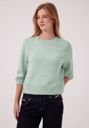 Ung kvinde med lysebrunt hår iført en lysegrøn trekvartærmer-sweater og mørke jeans, stående mod en ensfarvet baggrund.