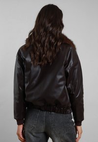 Giacca in ecopelle marrone con collo in pelliccia, vita elastica e polsini a coste. Modello indossa jeans grigi, vista da dietro.