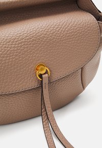 Brązowa skórzana torba z fakturą, z klapą i okrągłym zapięciem w złotym odcieniu oraz dwoma wąskimi skórzanymi frędzlami zwisającymi z zapięcia.