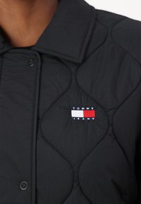 Chaqueta negra acolchada con un destacado parche del logo de Tommy Jeans en rojo, blanco y azul. Presenta un cuello con solapa y cierre a presión.