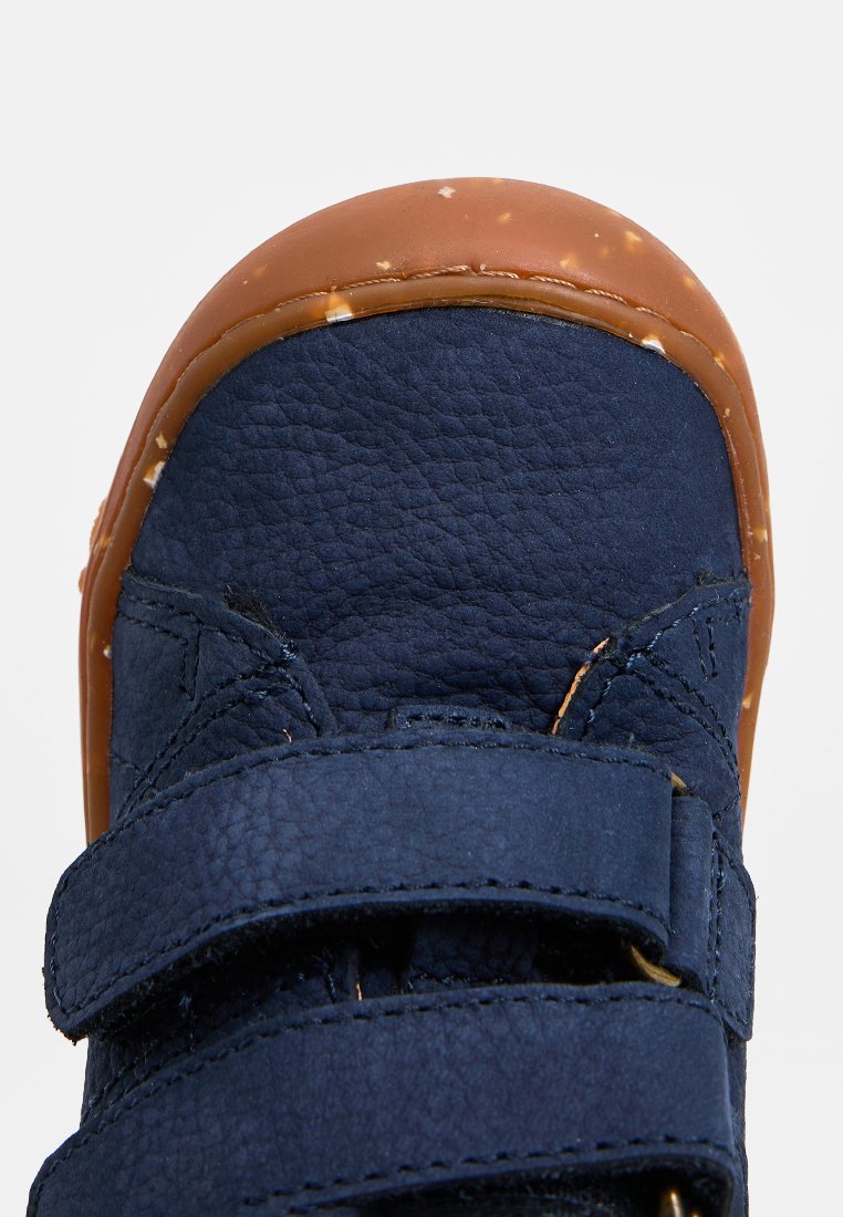 Marineblaue Lederschuhe mit einer abgerundeten braunen Gummisohle, ausgestattet mit zwei Klettverschlüssen und einem strukturierten Material im Oberteil für zusätzlichen Halt.