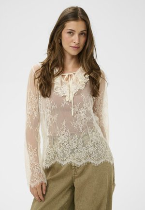 Jeune femme portant un chemisier en dentelle crème transparente à motifs floraux avec un lien au col, associé à un pantalon ample vert olive.