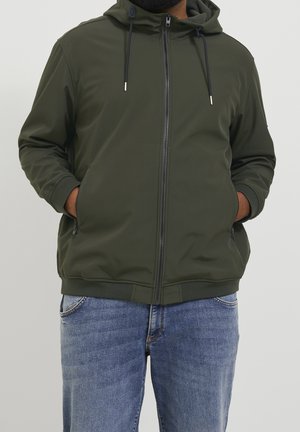 Homme portant une veste à capuche zippée vert foncé, mains dans les poches, et un jean bleu, devant un fond uni clair.